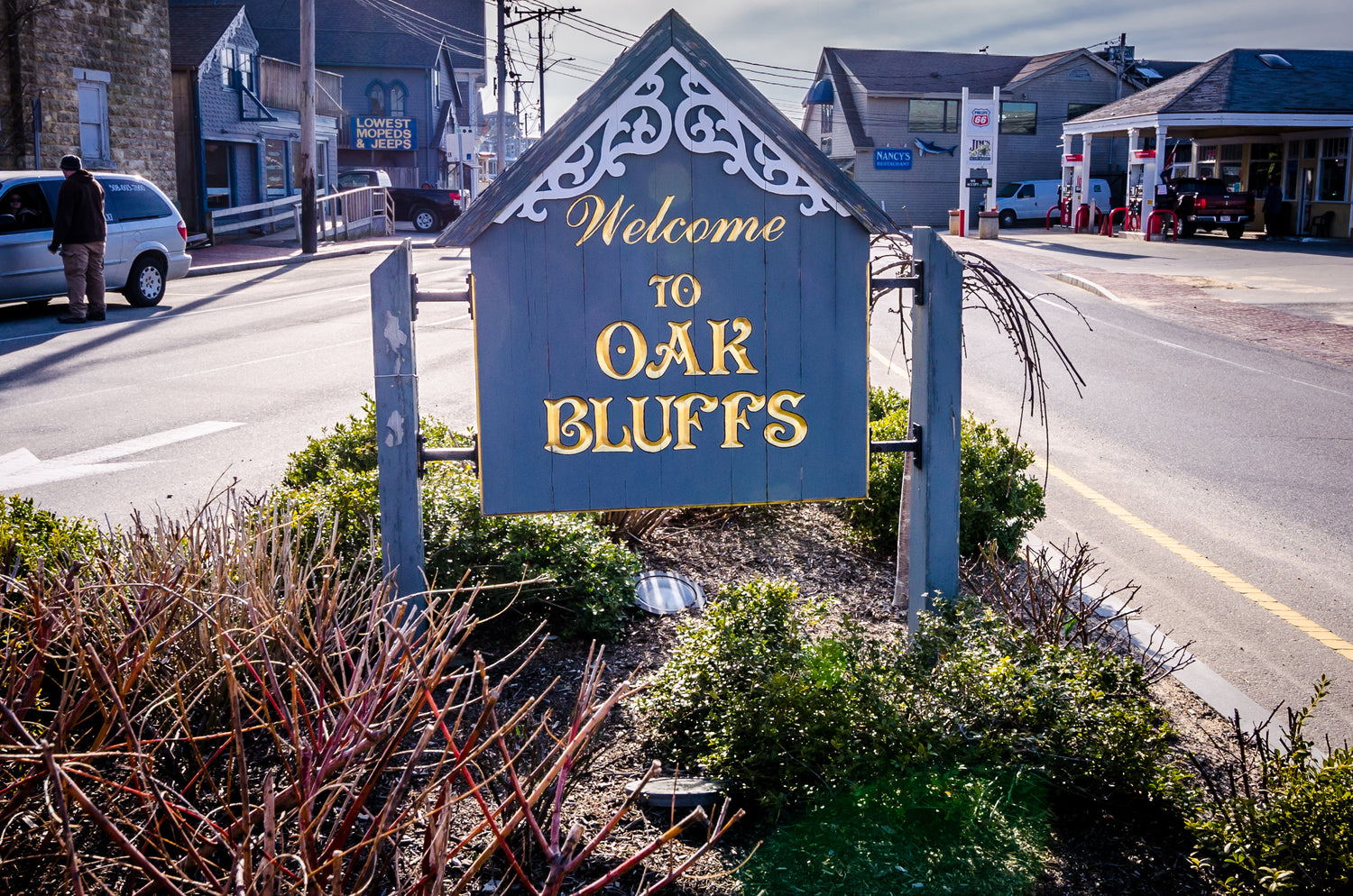 Welcome sign to Oak Bluffs with a street and buildings in the background
