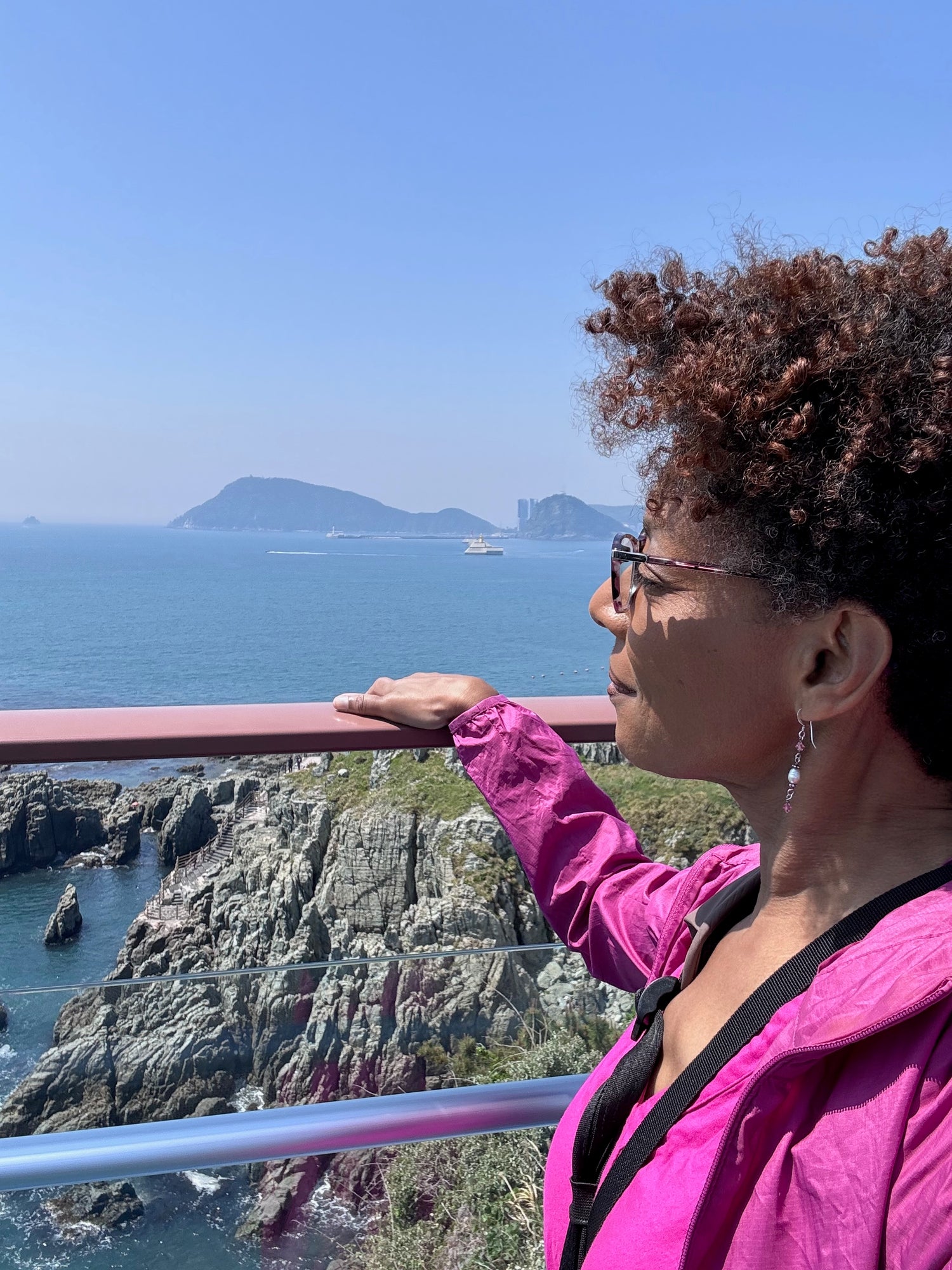 Asali Origins founder in a pink jacket looking at a scenic view of the ocean and cliffs.