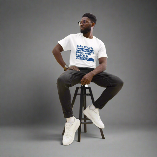 Man sitting on a stool wearing a white t-shirt with text, black pants, and white sneakers.